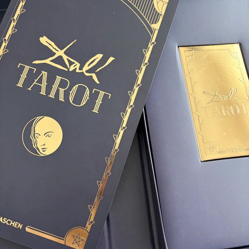 Guideboken till Dalí Tarot i mörkblått och guld bredvid den öppnade asken med kortlekens guldfärgade låda