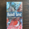 Asken till Haindl Tarot framifrån med collagelika målningar av ansikten och en röd måne i blått, rött och grått