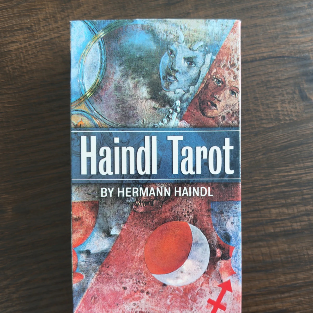 Asken till Haindl Tarot framifrån med collagelika målningar av ansikten och en röd måne i blått, rött och grått