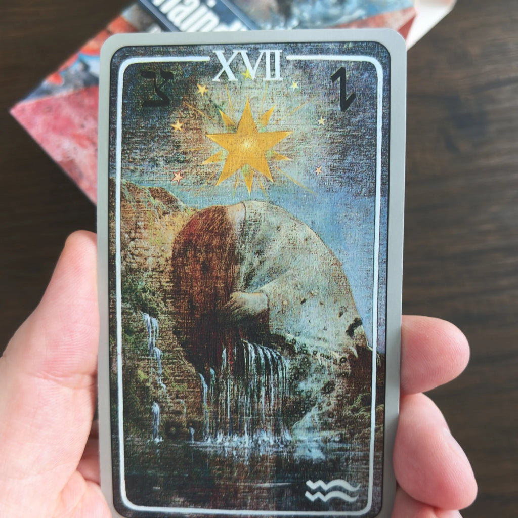 Kortet XVII The Star ur Haindl Tarot i en hand, med en gul stjärna över en böjd kvinna vid ett vattenfall