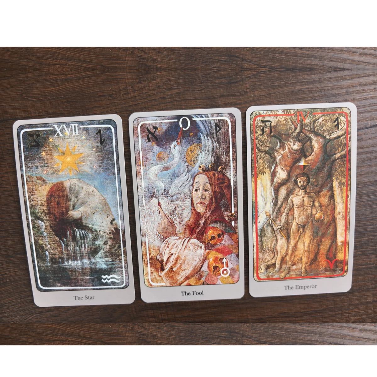 Tre kort ur Haindl Tarot bredvid varandra på ett träbord: The Star, The Fool och The Emperor