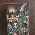 Asken till The Lord of the Rings Tarot Deck & Guide framifrån med träsnittsliknande teckning av en trollkarl och en hobbit