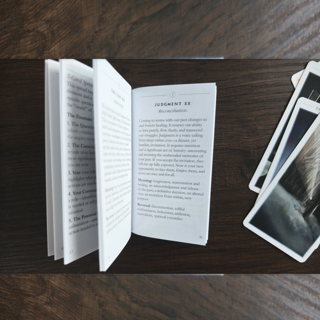 Uppslag i guideboken till The Fountain Tarot med texten om Judgment XX, bredvid några kort på träbordet