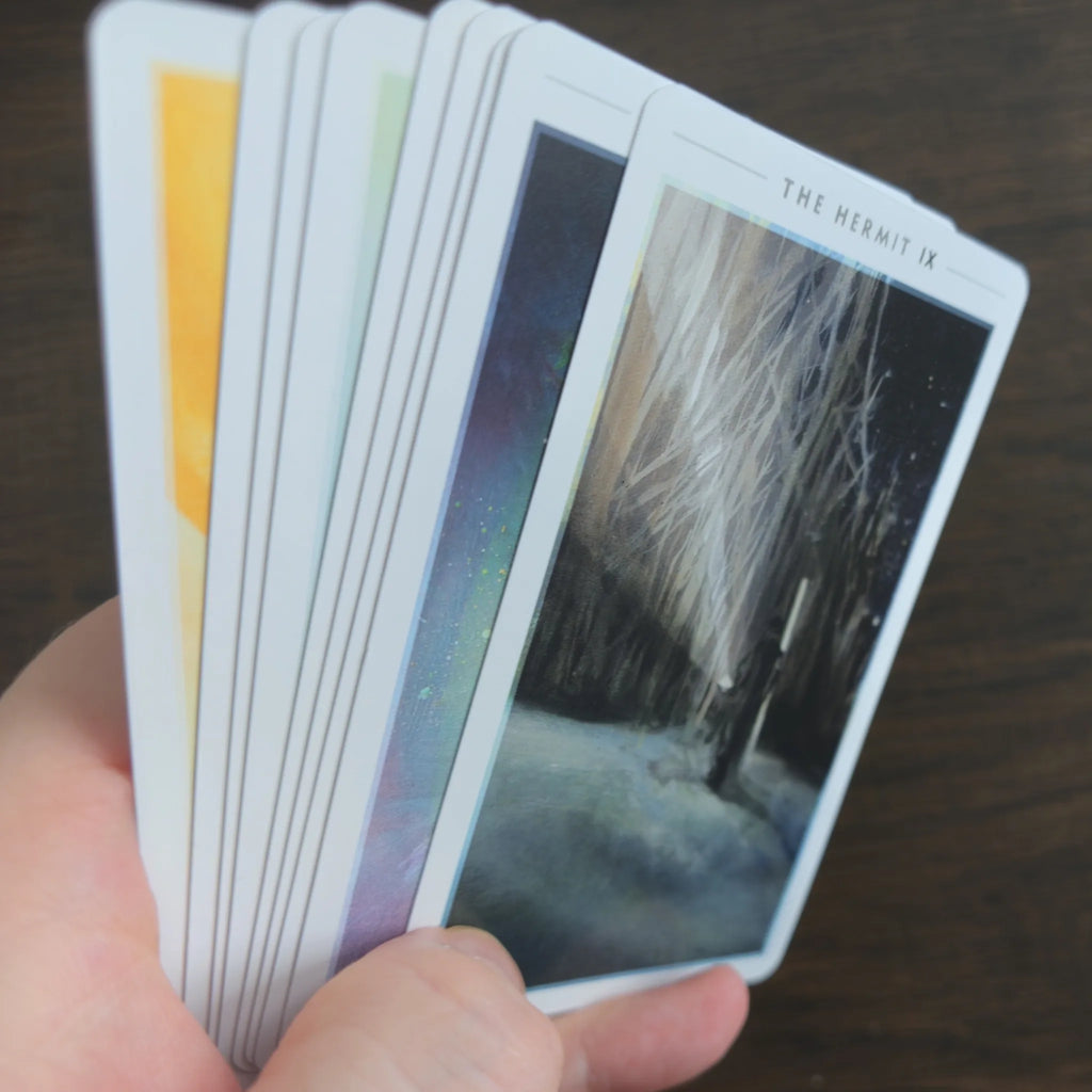En hand håller en solfjäder av kort ur The Fountain Tarot med The Hermit överst i snöigt nattlandskap