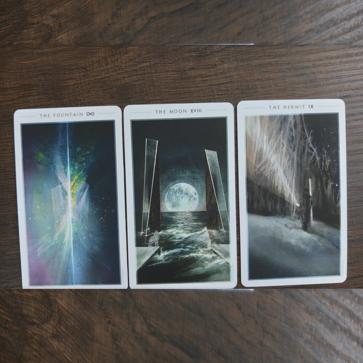 Tre kort ur The Fountain Tarot bredvid varandra på ett träbord: The Fountain, The Moon och The Hermit
